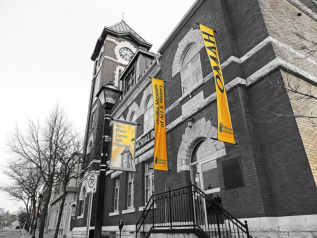 Deb Halbot OMAH building Historic brick building with clock tower, arched doorway, steps and railing, bare trees, and tall yellow banners reading "OMAH" and "Orillia Museum of Art & History".